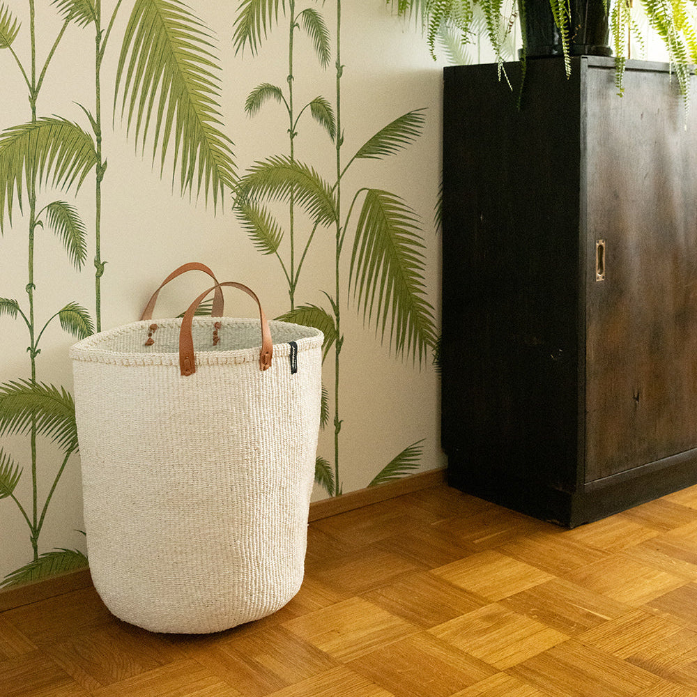 White woven basket with brown handles on a wooden floor against a wall with green leafy pattern.