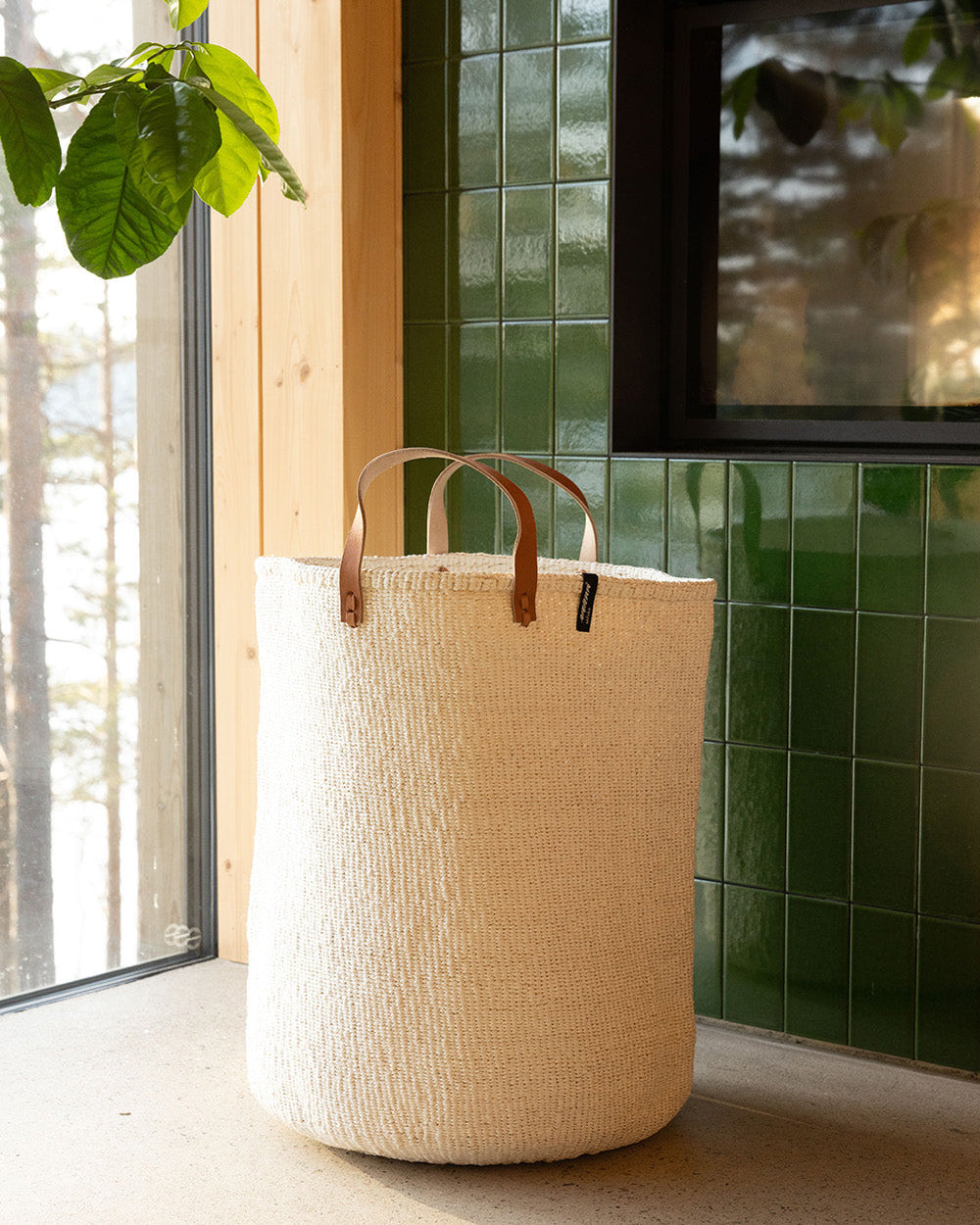 Beige woven basket with brown handles on a light wooden floor against a green tiled wall.