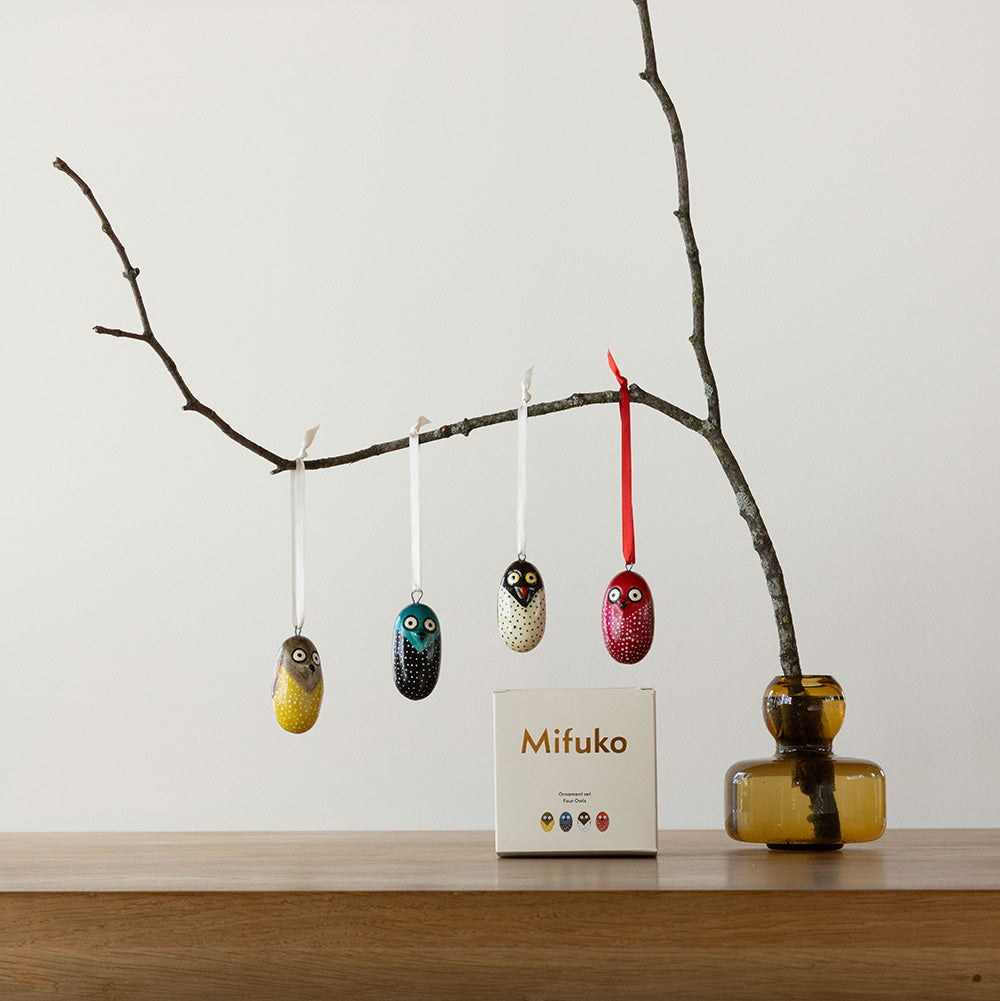 Decorative owl ornaments hanging from a branch with a vase on a wooden surface.
