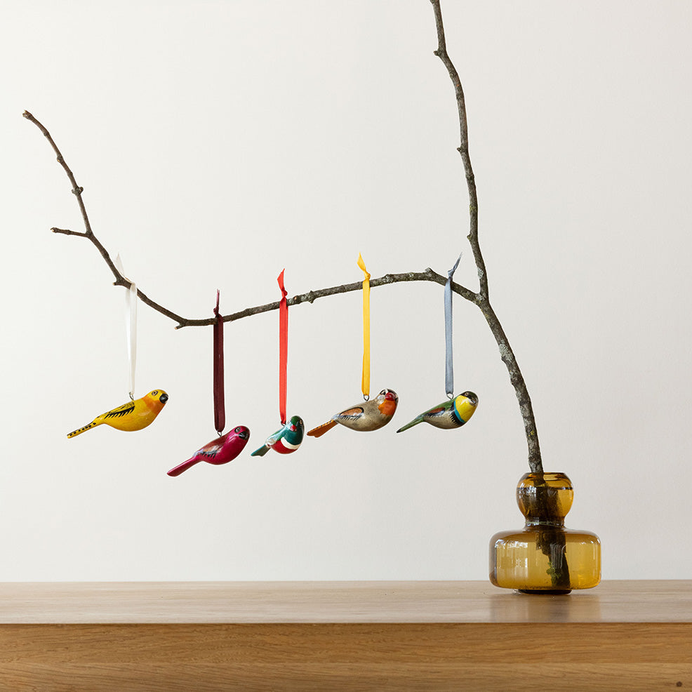 Decorative birds hanging from a branch with a yellow vase on a wooden surface.