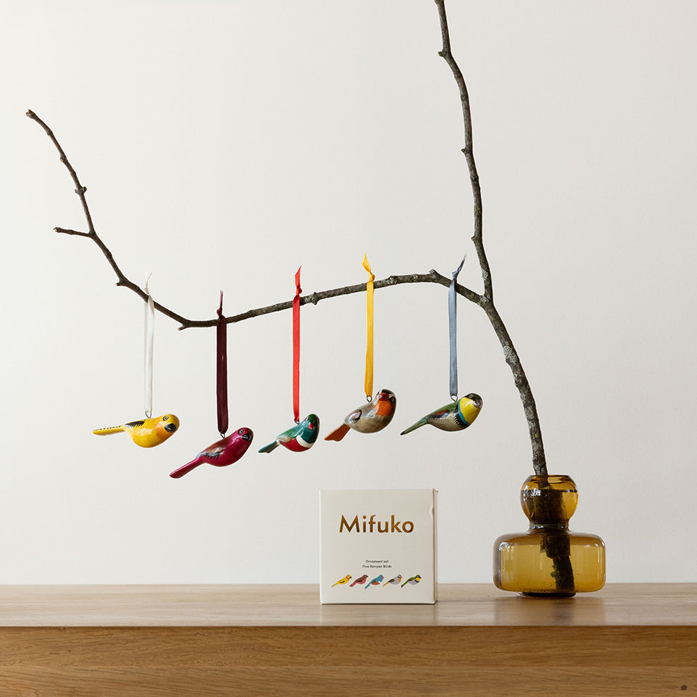 Colorful bird-shaped ornaments hanging from a branch with a 'Mifuko' box on a wooden surface.