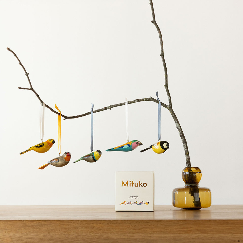 Decorative birds hanging from a branch with a Mifuko product box on a wooden surface.