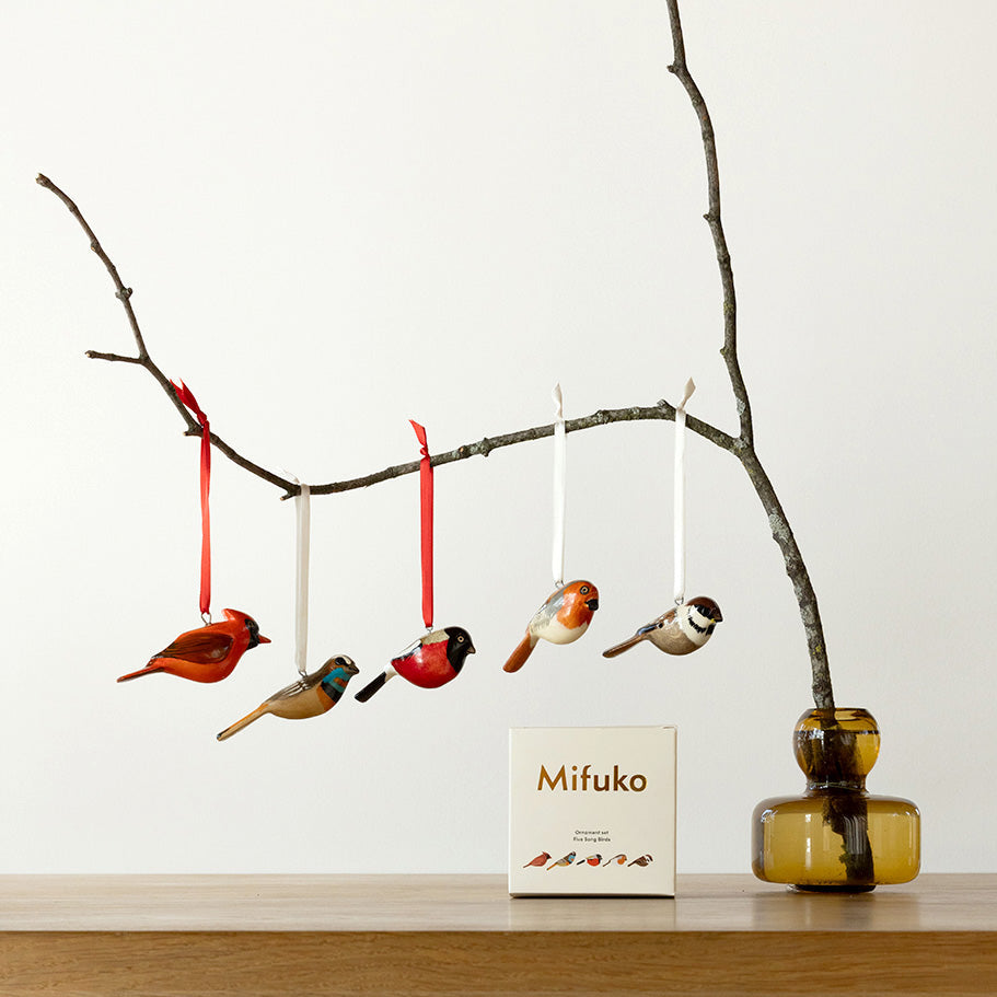 Decorative bird ornaments hanging from a branch with a Mifuko box on a wooden surface.