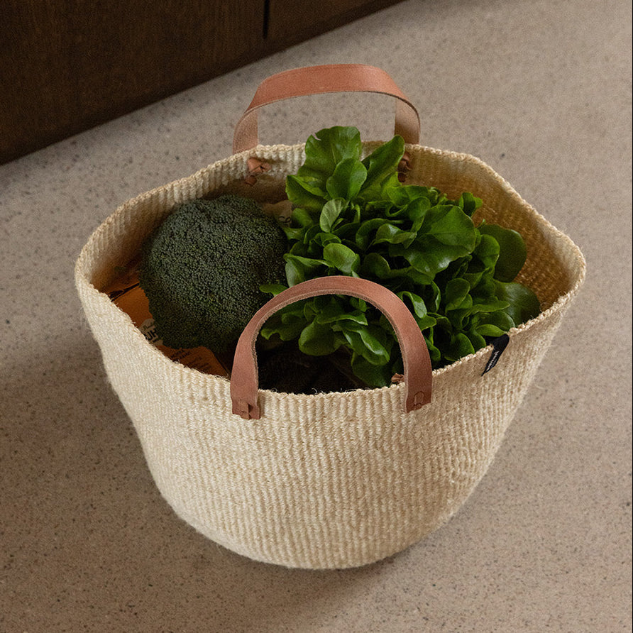 Woven basket with handles containing plants on a beige floor.