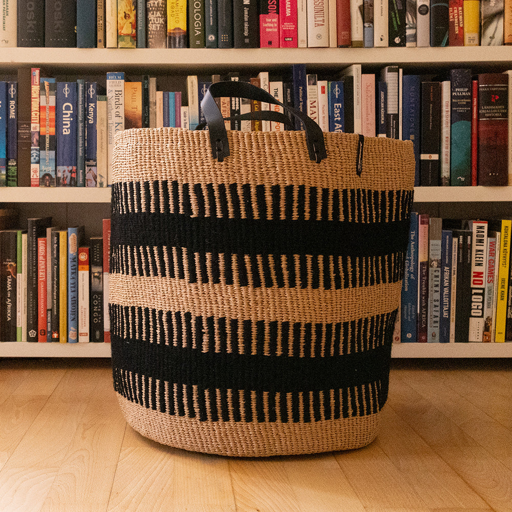 Woven basket with black stripes on a wooden floor in front of a bookshelf filled with books.