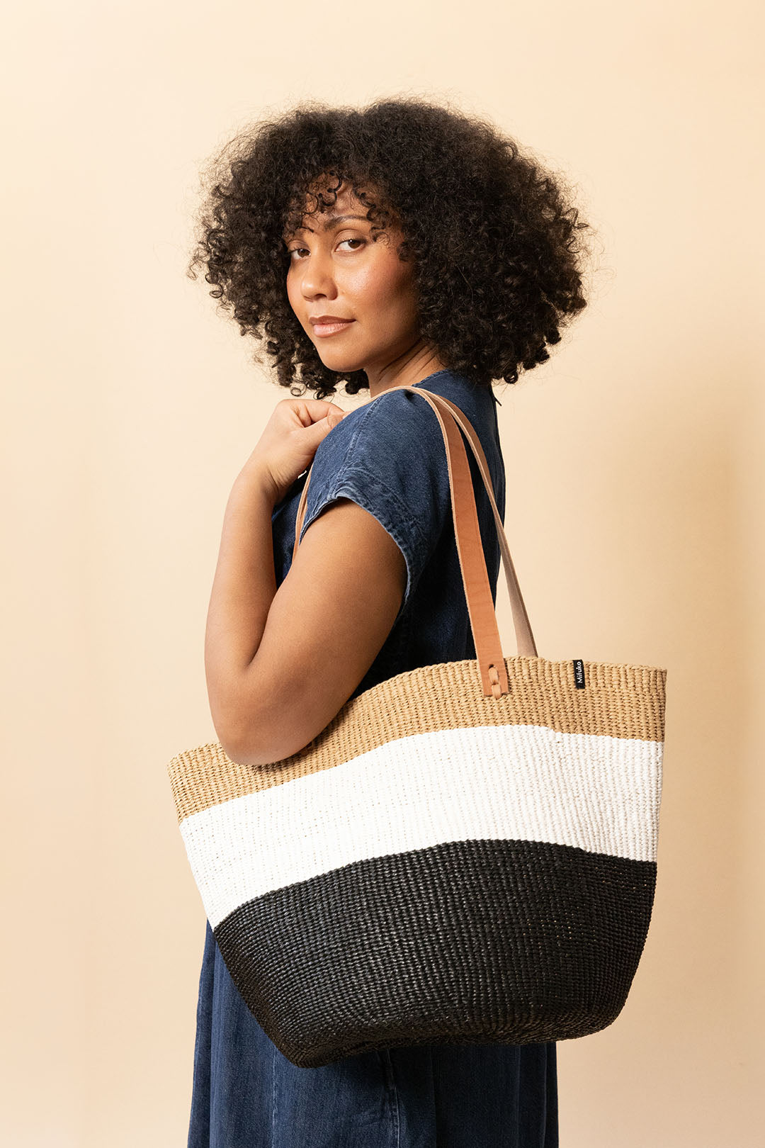 A woman with curly black hair wearing a blue denim dress carries a large woven tote bag. The bag features three horizontal color-blocked stripes: a natural beige top stripe, a wide white middle stripe, and a black bottom stripe, with tan leather shoulder straps.