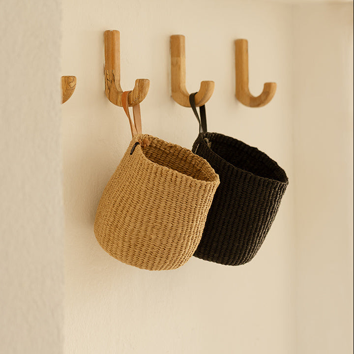 Two woven baskets, one brown and one black, hanging from wooden hooks on a beige wall.