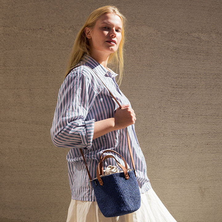 A woman wearing a blue and white striped button-up shirt and a cream skirt carries a small navy blue woven handbag with brown leather handles against a textured beige wall.