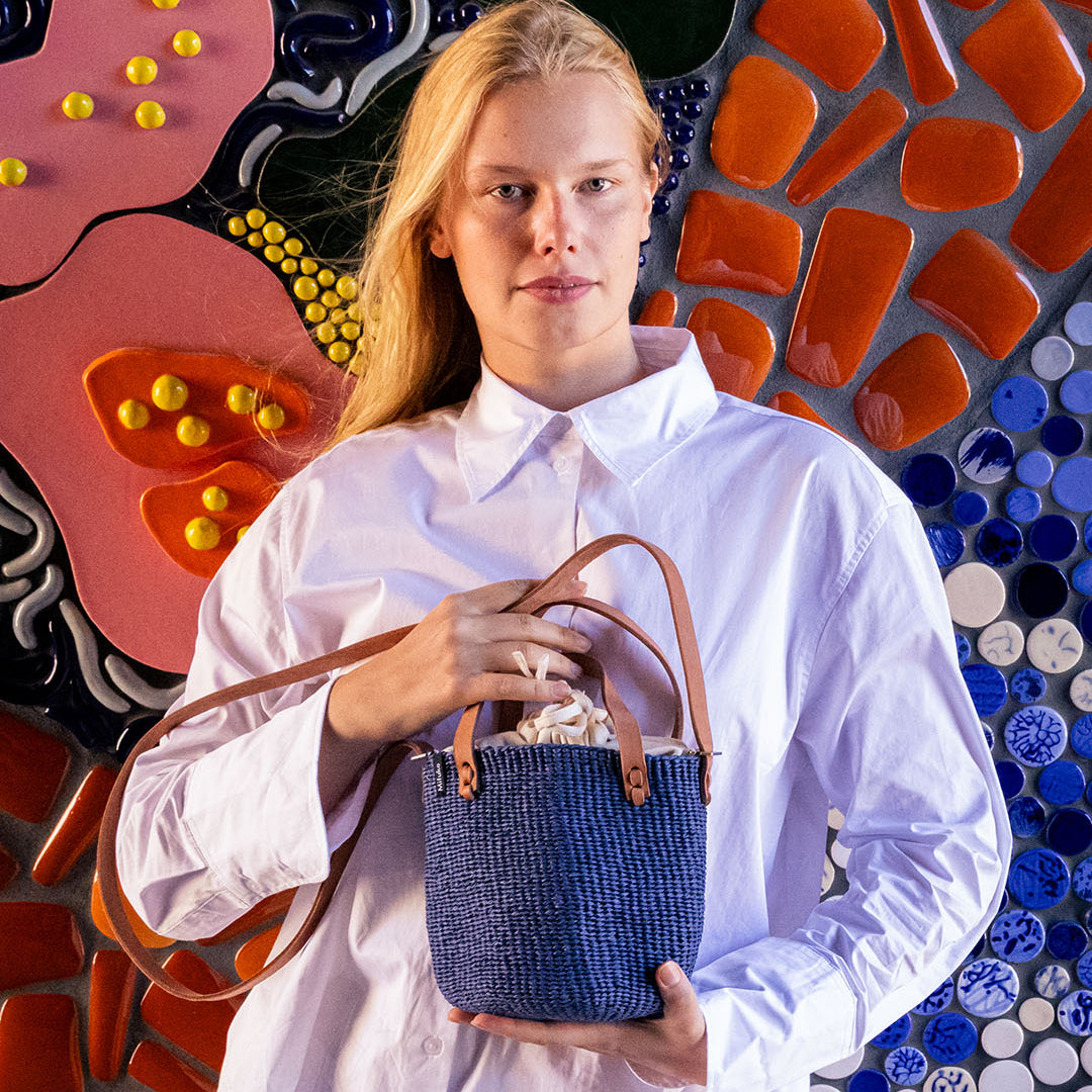 A woman wearing a white button-down shirt and cream skirt holds a small blue woven basket bag with brown leather handles in front of a colorful mosaic wall.