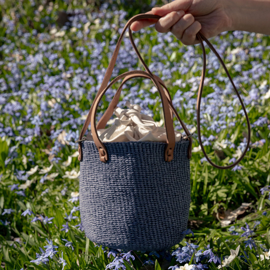 A woven blue basket bag with tan leather handles is held by a person's hand in a grassy field filled with small blue flowers.