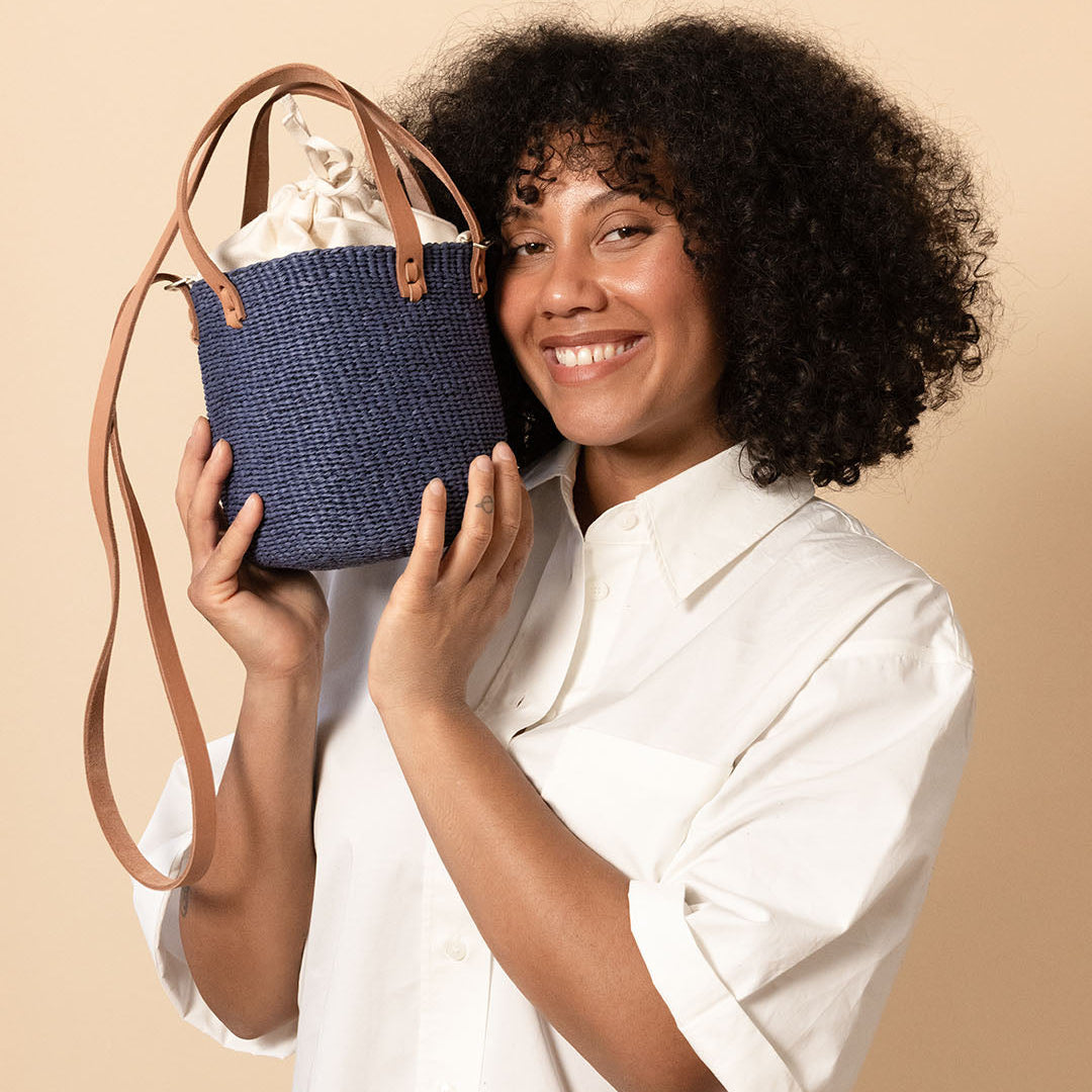 A smiling woman with curly dark hair holds a small woven blue bag with tan handles and a long strap against a plain beige studio background.