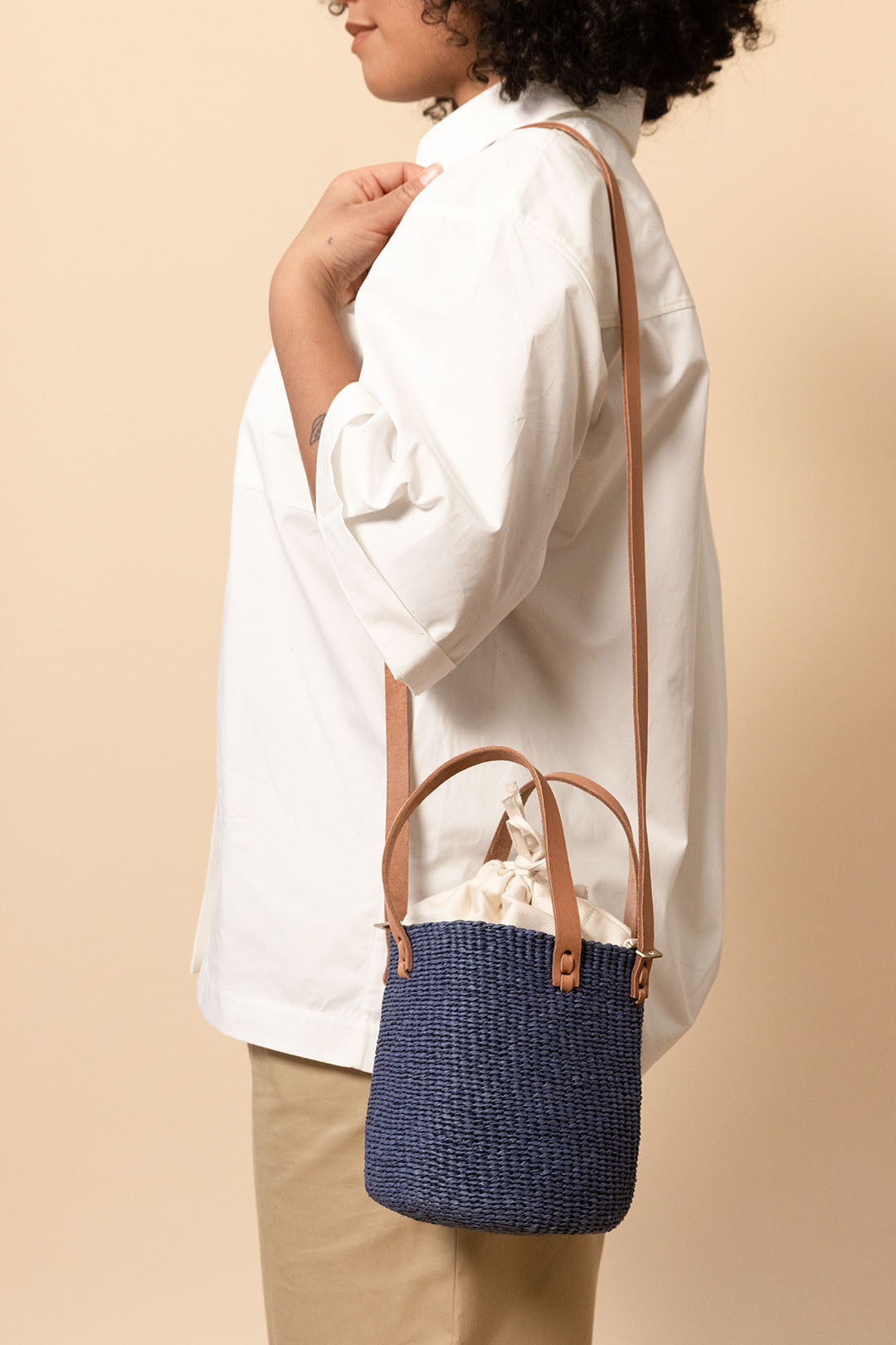 A person wearing a white button-up shirt and beige pants carries a small woven bucket-style shoulder bag in navy blue with tan leather handles and strap against a plain beige studio background.