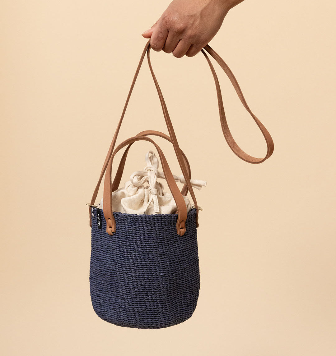 A navy blue woven bucket bag with tan leather handles and a drawstring detail, held by a person's hand against a solid beige background.