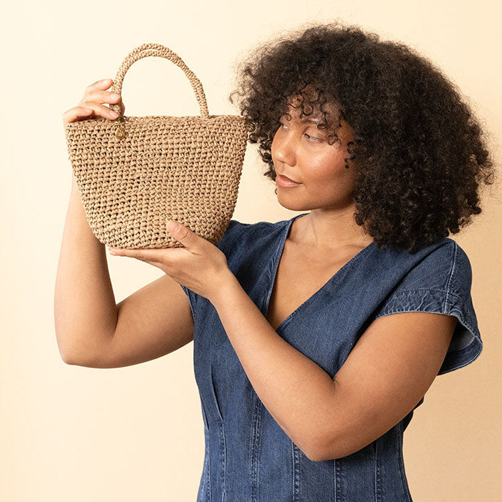 A woman with curly dark hair wearing a blue denim dress holds up a woven straw tote bag against a solid beige background.