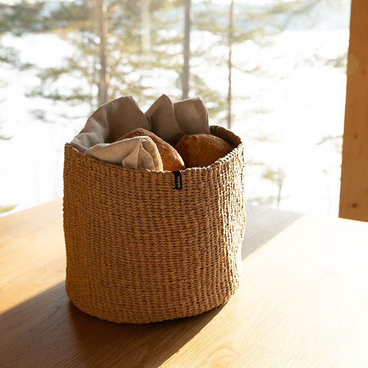 Woven basket with bread on a wooden surface near a window with a view of trees.