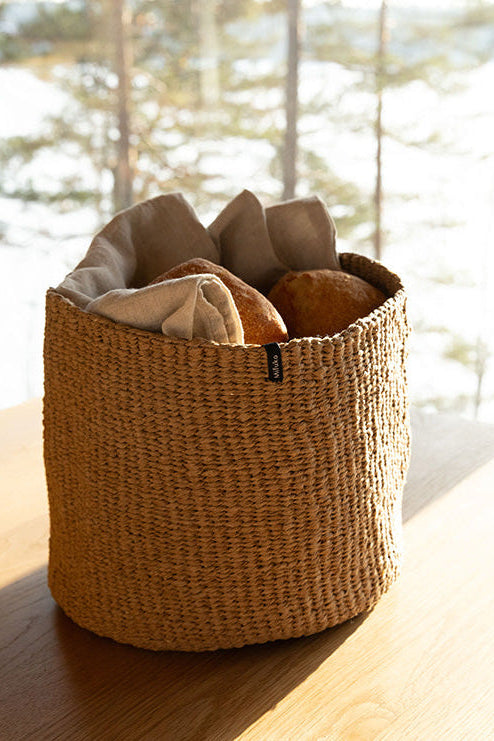 Woven basket with bread on a wooden surface near a window with a view of trees.