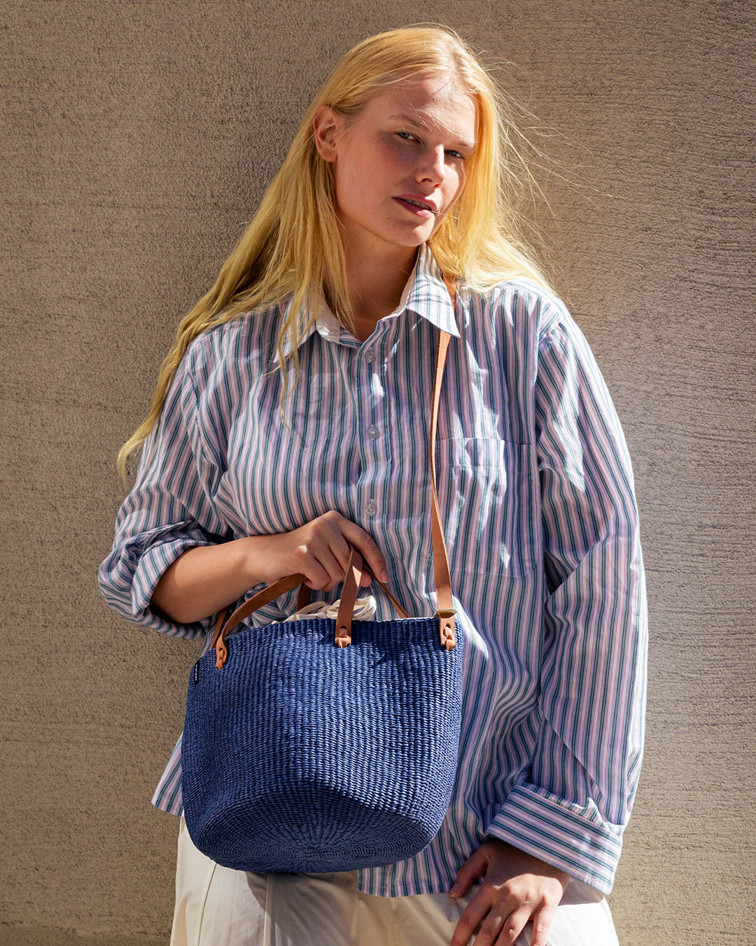 A woman stands in front of a wall, wearing a white and blue striped shirt and holding a navy blue woven shoulder bag.