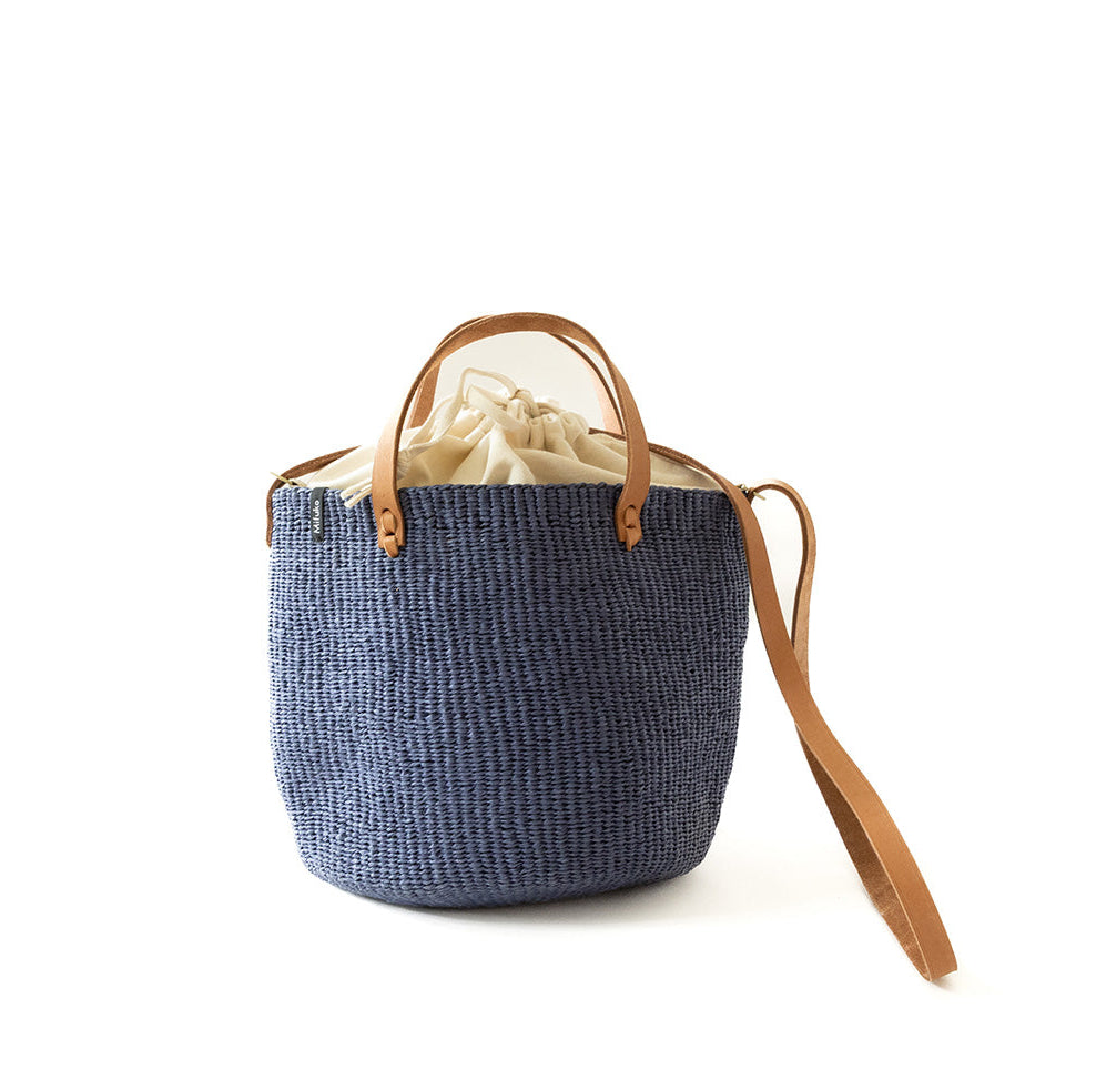 A navy blue woven straw bucket bag with tan leather handles and a matching shoulder strap, featuring a cream-colored drawstring closure at the top, set against a plain white background.