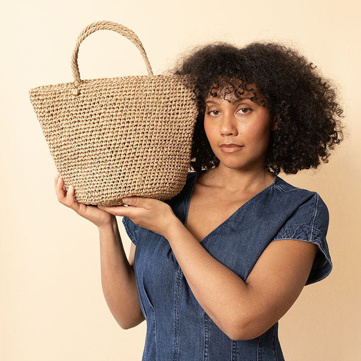 A woman with curly dark hair wearing a blue denim dress holds a woven straw tote bag against a solid beige background.