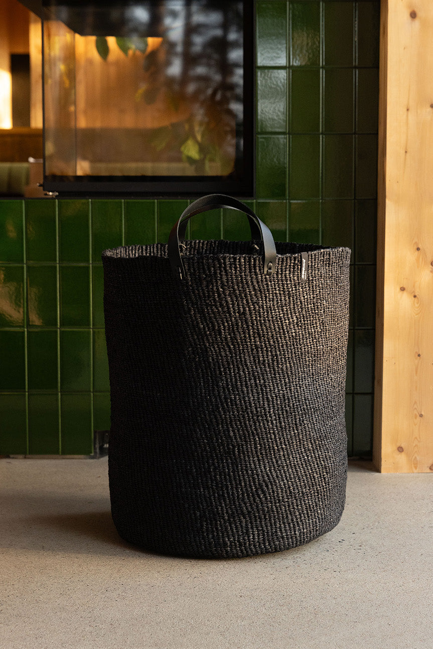 Black woven basket on a gray surface with a green tiled wall and wooden panel in the background.