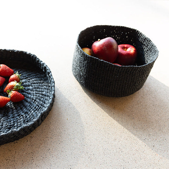 Two black woven bowls with strawberries and apples on a light surface