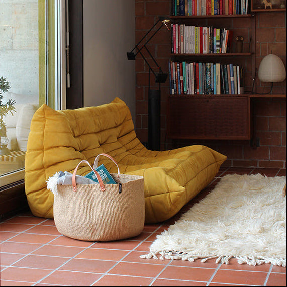 Living room with yellow sofa, handwoven brown basket, bookshelf, and brick wall.