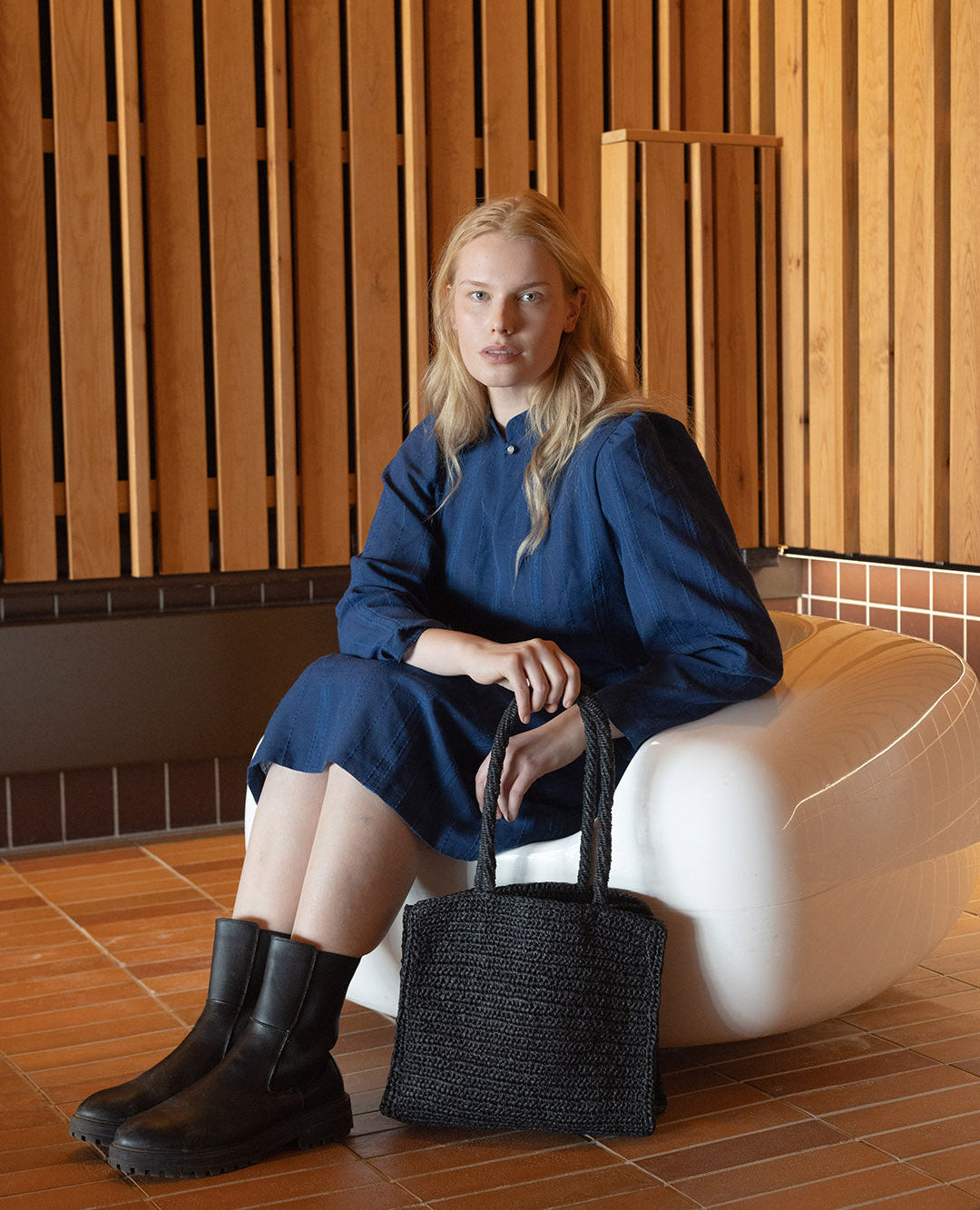Woman in a blue dress sitting on a white chair holding a black handbag in a room with wooden walls.