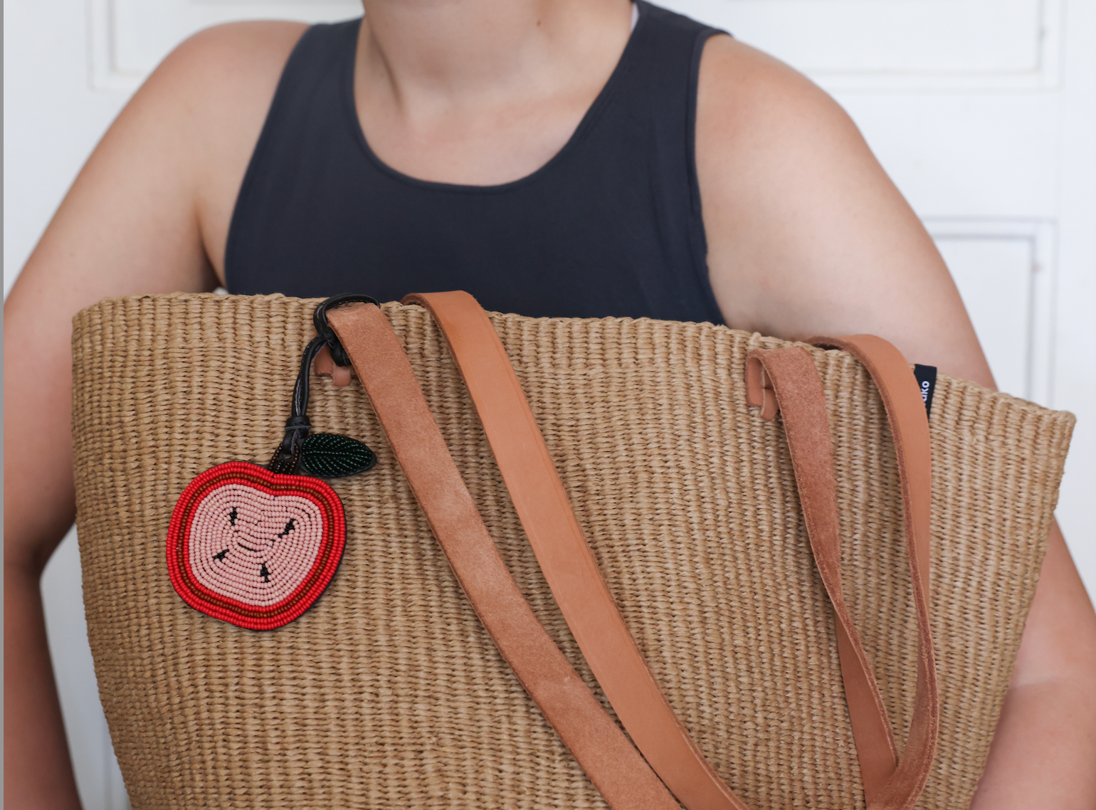 A woman wearing a black tank top holds a large woven straw tote bag with tan leather shoulder straps and a red embroidered apple patch on the front, set against a white paneled wall.