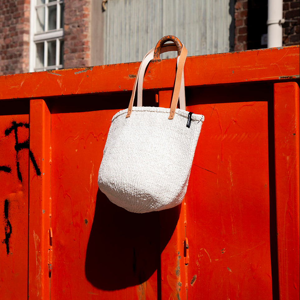 White handbag with brown strap hanging on a red metal fence with graffiti.