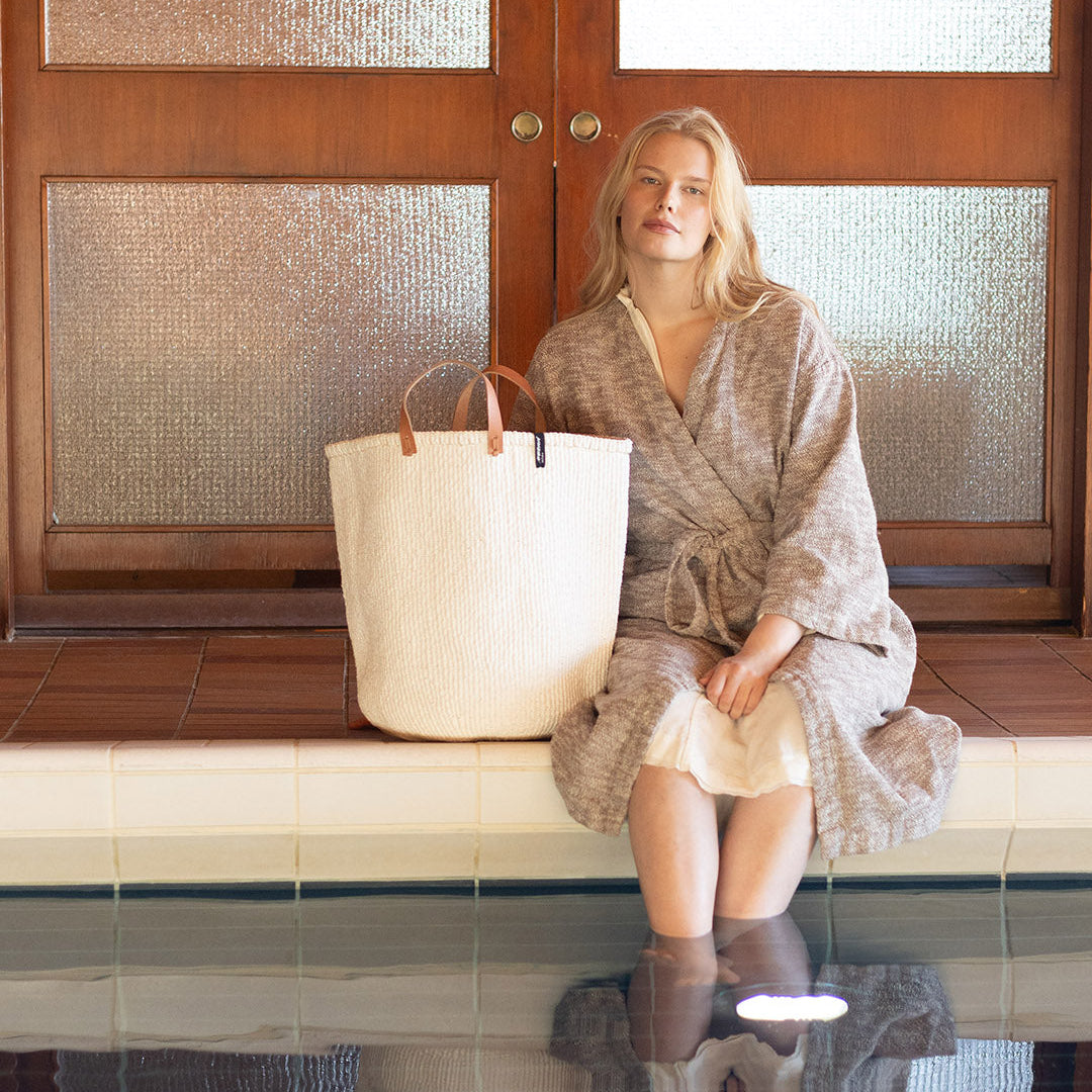Woman in a robe sitting by a pool with a large bag next to her.