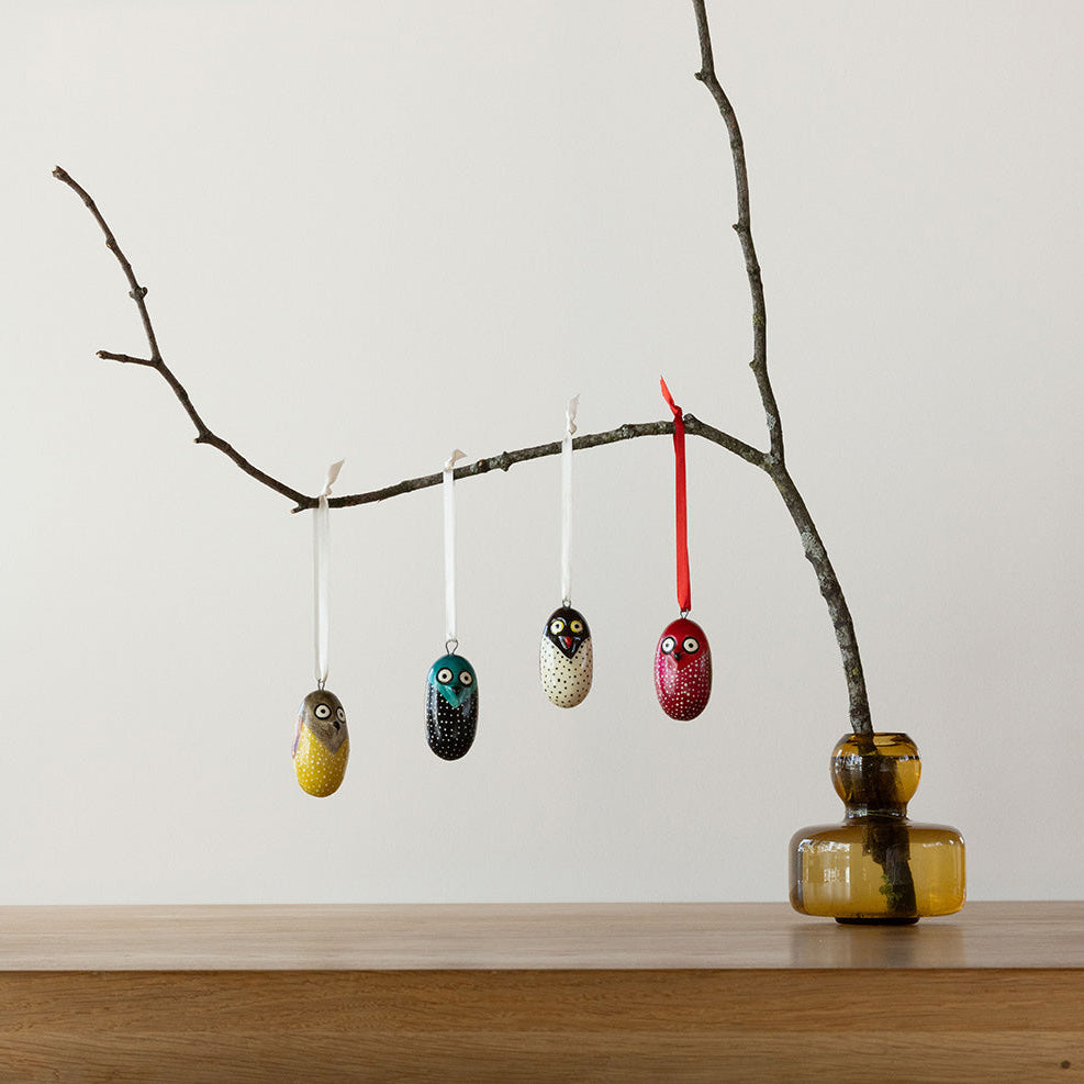 Decorative owl ornaments hanging from a branch with a vase on a wooden surface.