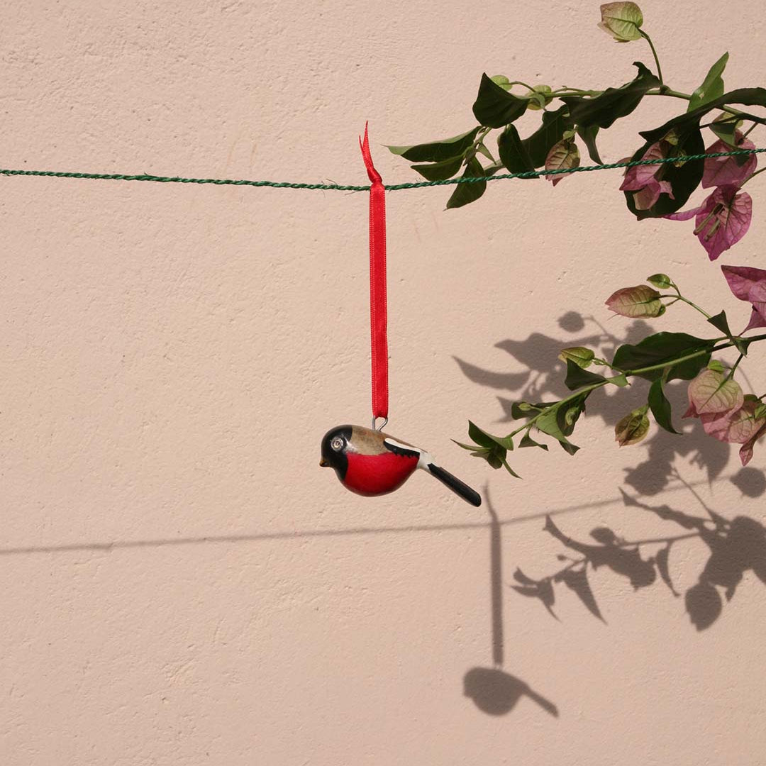 Decorative bird ornament hanging from a red ribbon against a beige wall with greenery.