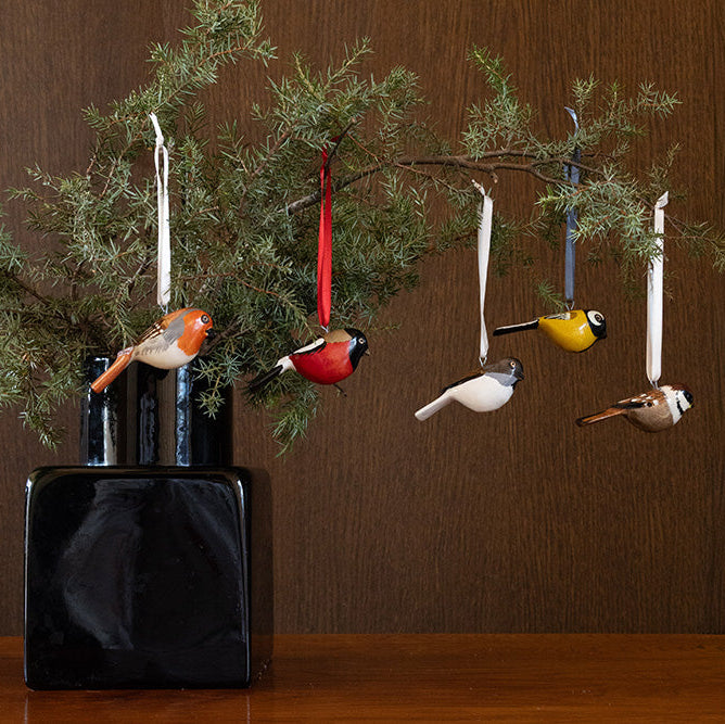 Decorative bird ornaments hanging from a branch in front of a dark vase on a wooden surface.
