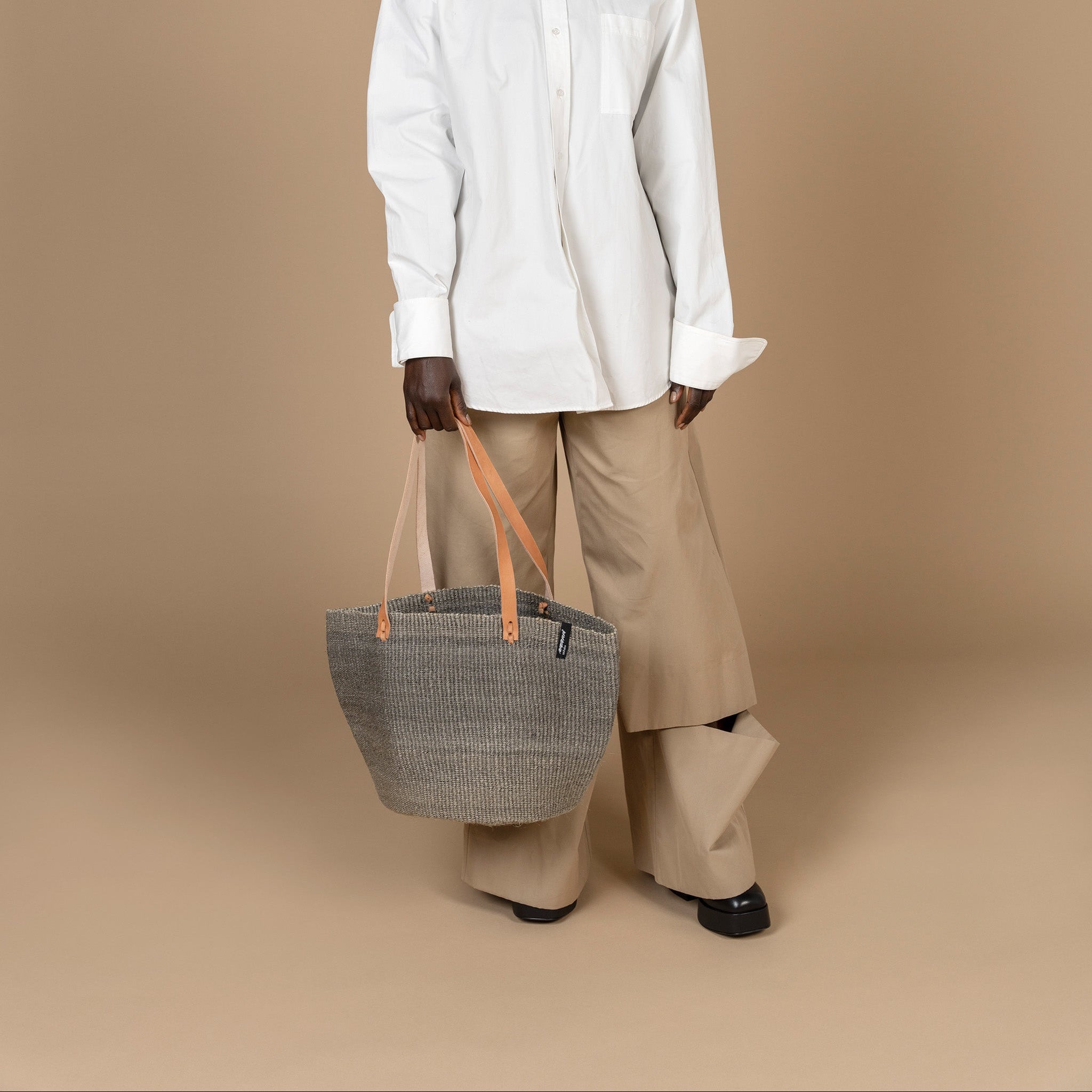 Person wearing a white shirt and beige pants holding a gray bag against a beige background