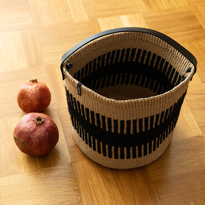 A woven basket with black and beige horizontal stripes and a black handle, placed on a light wood parquet floor next to two pomegranates.