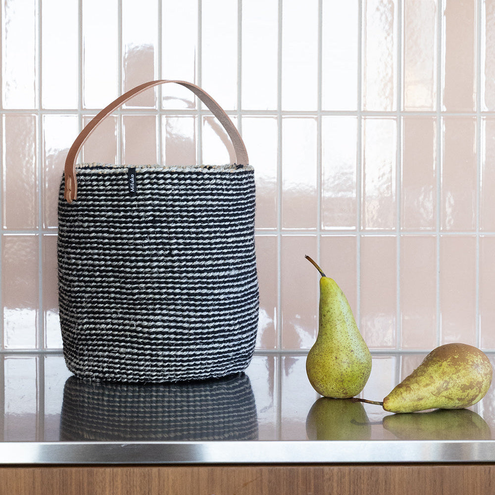 A black and white woven basket bag with a tan leather handle sits on a stainless steel kitchen counter next to two green pears, set against a tiled backsplash.