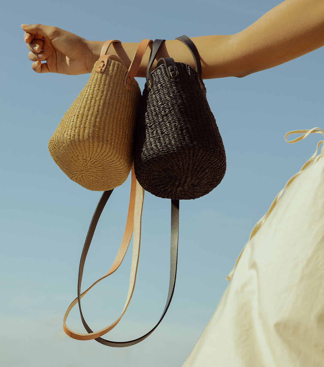 Two woven handbags, one beige and one black, held by a person against a clear blue sky.