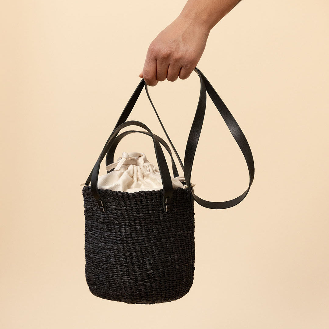 A hand holding a small black woven bucket bag with black shoulder straps and a cream drawstring closure, set against a solid beige background.