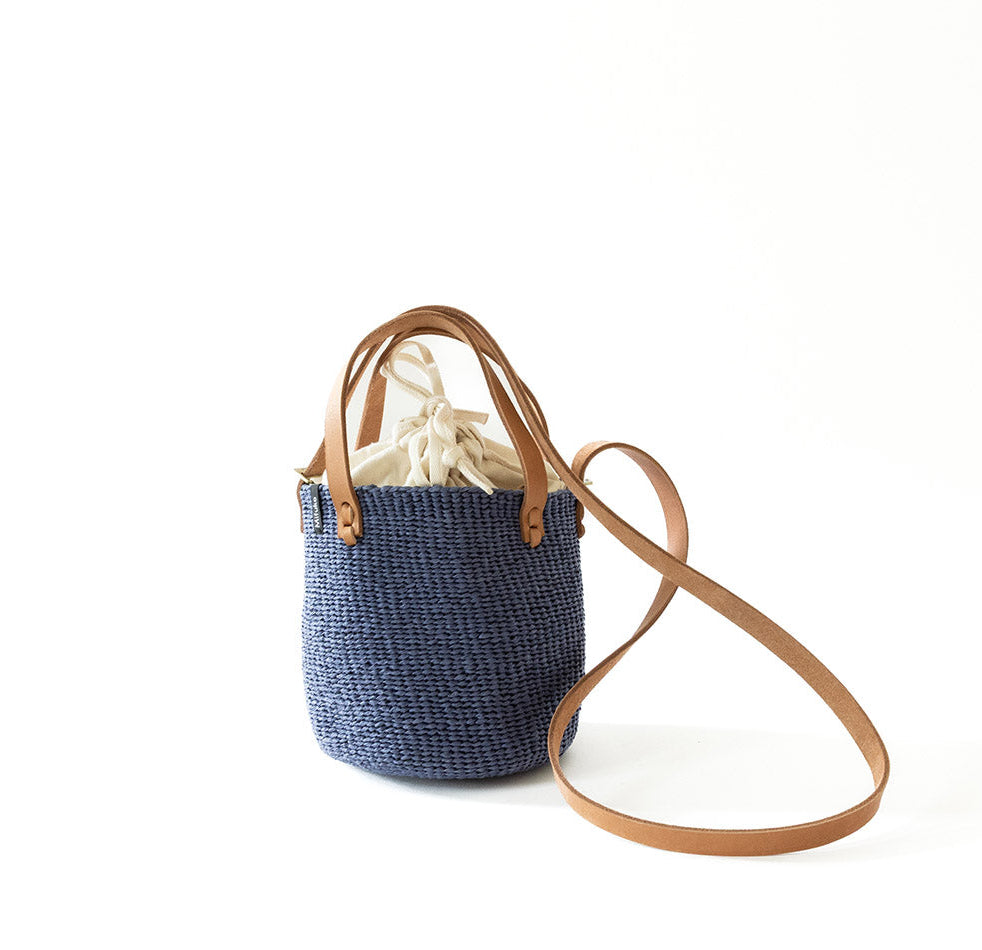 A navy blue woven straw bucket bag with tan leather handles and a matching shoulder strap, shown on a clean white background.
