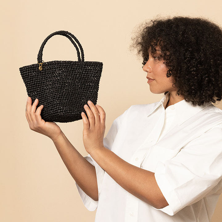 A woman with curly dark hair wearing a white button-up shirt holds up a small black woven handbag against a solid beige background.