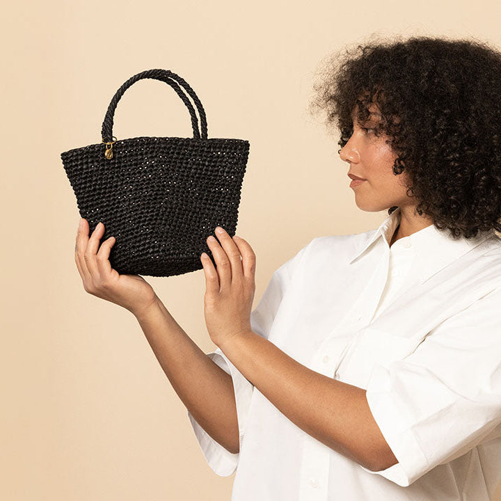 A woman with curly dark hair wearing a white button-up shirt holds up a small black woven handbag against a solid beige background.