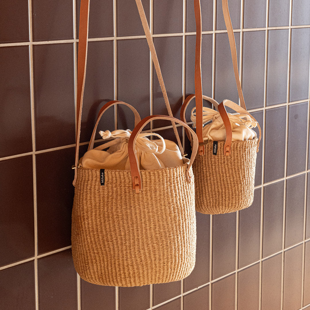 Two woven bags with cotton inner pouches and leather straps hanging on a tiled wall.