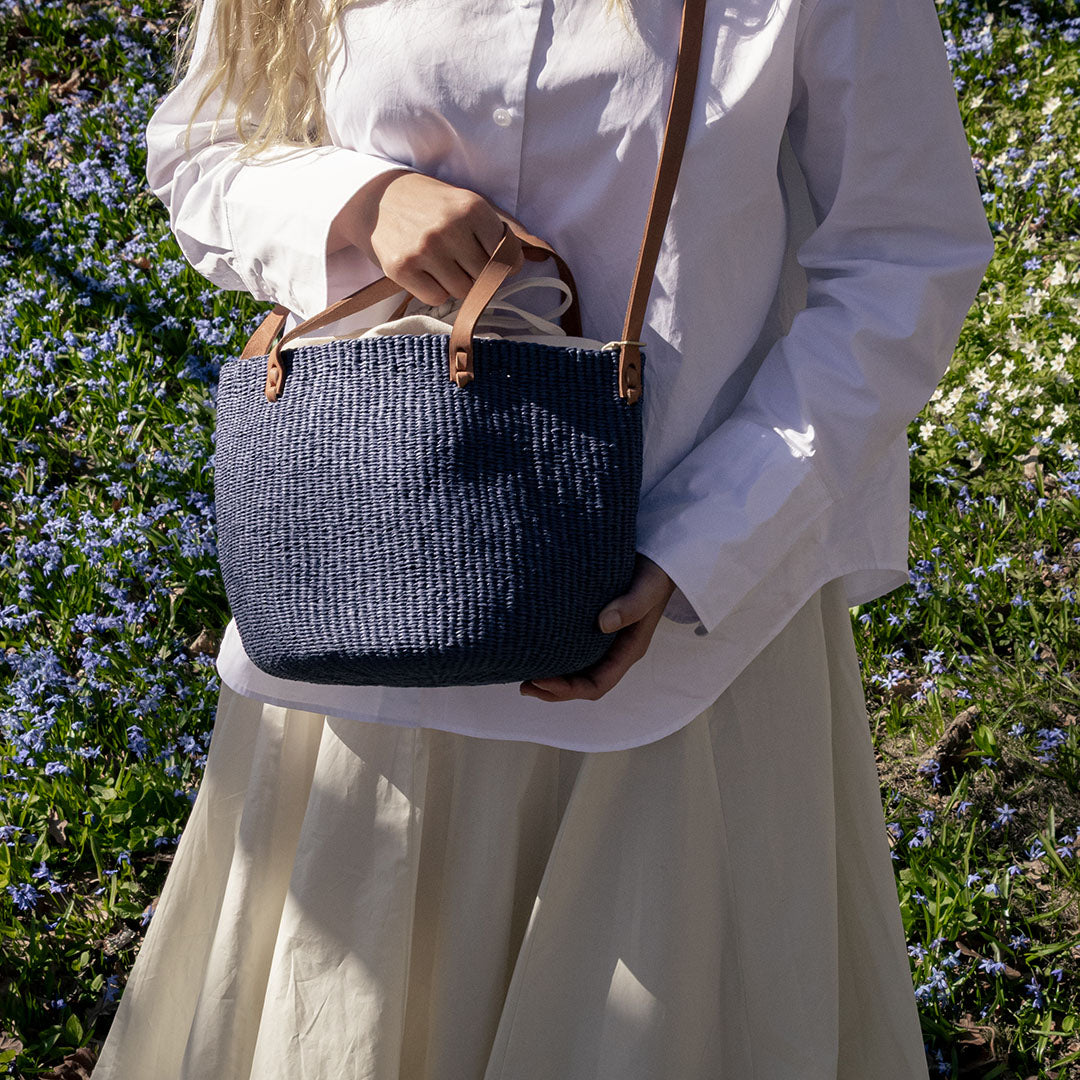A person in a white blouse and cream skirt holds a navy blue woven straw tote bag with tan leather handles and strap in an outdoor garden setting with purple flowers.