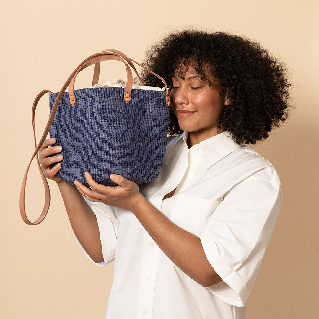 A woman with curly black hair wearing a white button-up shirt and beige pants holds a blue woven tote bag with tan leather handles against a solid beige background.