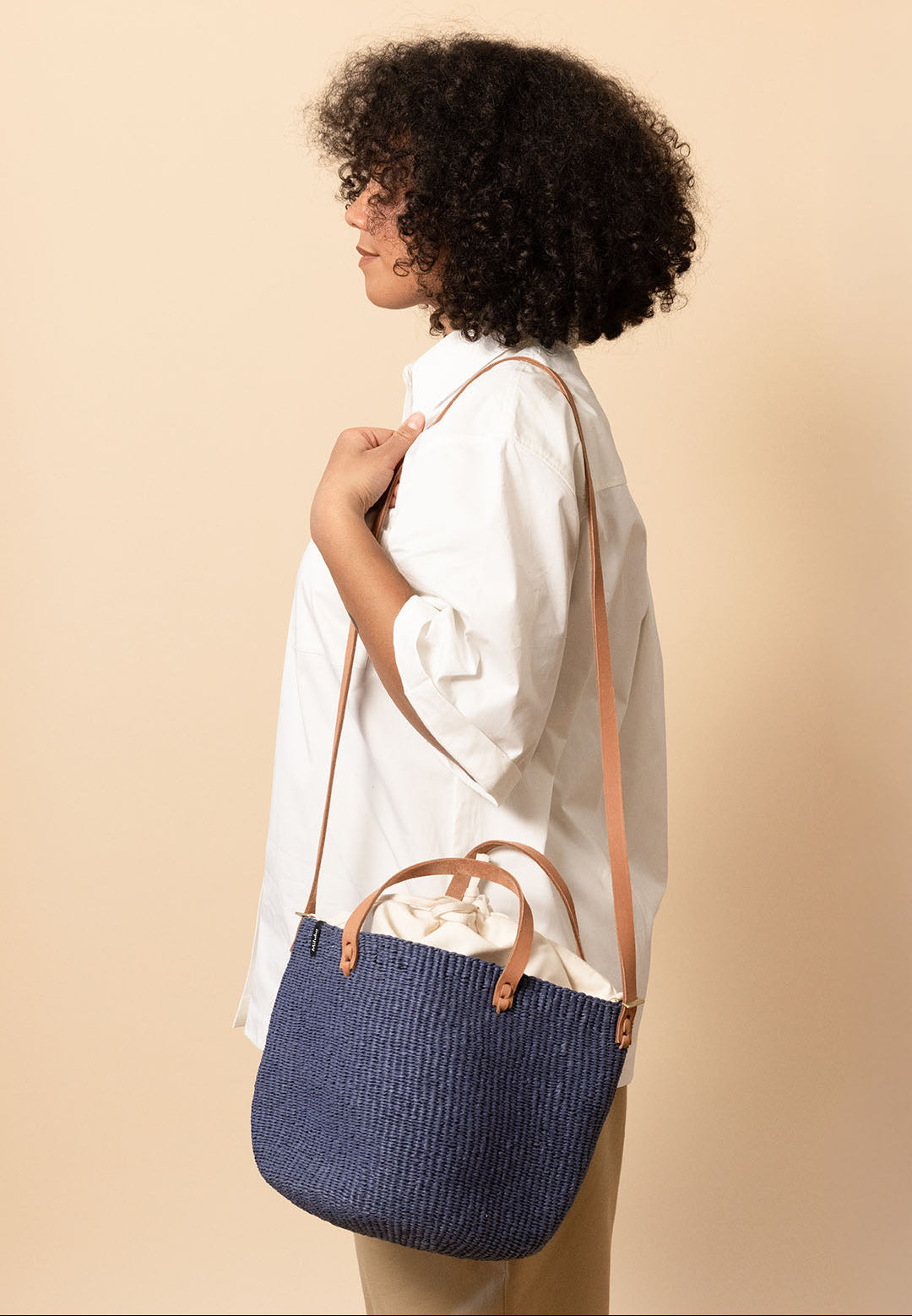 A woman with curly black hair, wearing a white shirt and beige pants, shown in profile carrying a blue woven straw tote bag with tan leather handles and strap against a solid beige background.