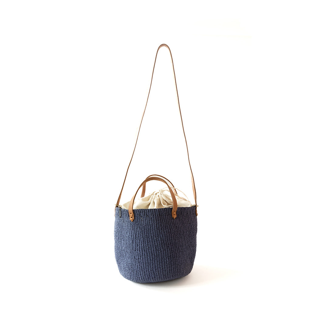 A navy blue woven bucket bag with a long tan shoulder strap and a cream drawstring closure at the top, shown on a plain white background.