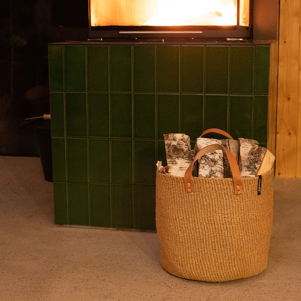 Wicker basket with books in front of a lit fireplace with green tiles.