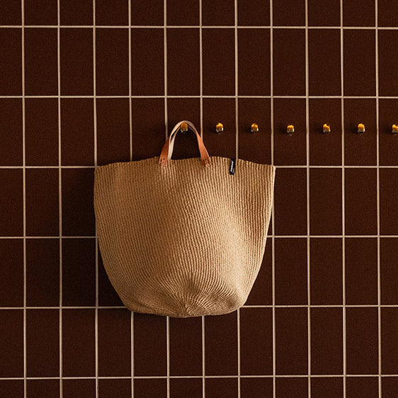 Beige woven bag hanging on a dark brown grid wall.