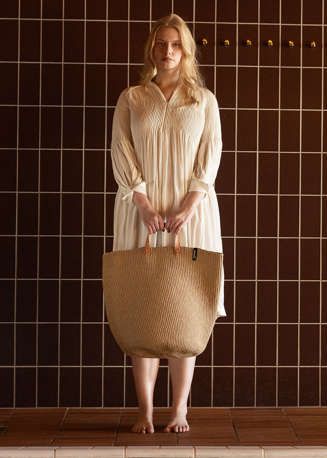 Woman holding a woven bag against a tiled wall.