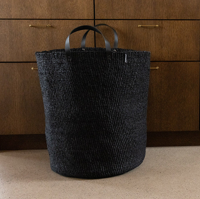 Black woven basket with leather handles on a beige floor against wooden cabinets.